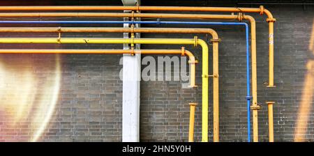 Pipes in yellow, orange and blue bend at right angles in front of a brick wall, with light beams Stock Photo