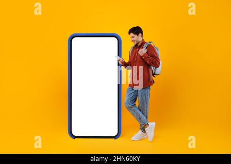Traveler Guy Using Phone Standing Near Big Cellphone, Yellow Background Stock Photo