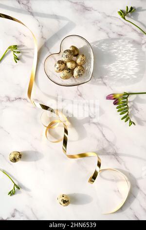 Golden quail eggs in glass heart shaped bowl. Twigs with young leaves on off white marble. Spring, Easter off white background with purple freesia Stock Photo