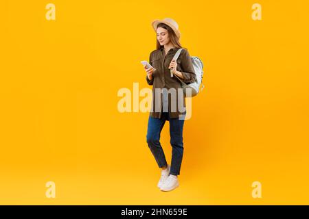 Tourist Lady Using Smartphone Browsing Internet Holding Backpack, Yellow Background Stock Photo