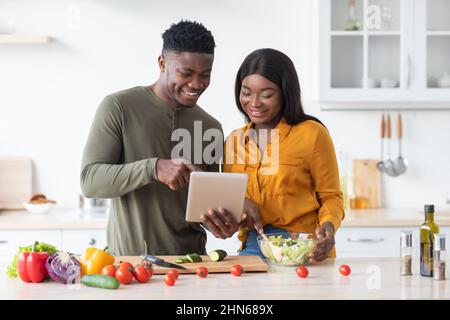 Online Recipe. Happy Black Spouses Using Digital Tablet In Kitchen While Cooking Stock Photo