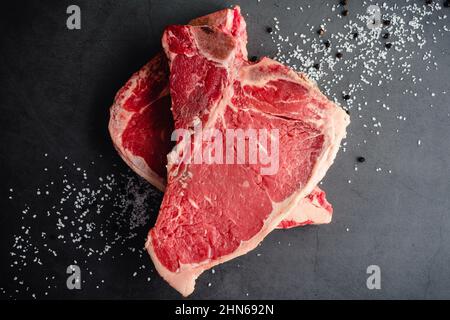 Raw T-Bone Steaks with Kosher Salt and Black Peppercorns: Uncooked bone-in beef steaks with salt and pepper on a dark background Stock Photo