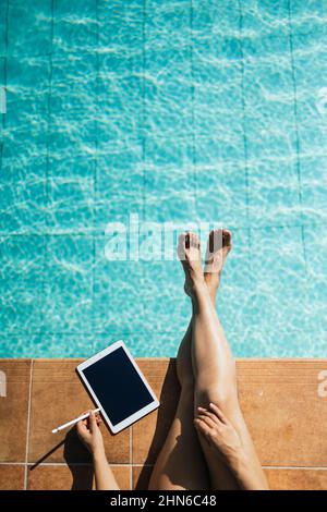 Unrecognizable woman using a tablet at the edge of a turquoise pool in portrait format Stock Photo