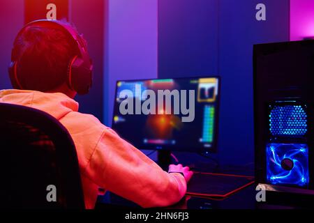 Back view of teenager boy play computer video game in dark room, use neon colored rgb mechanical keyboard, place for cybersport gaming Stock Photo