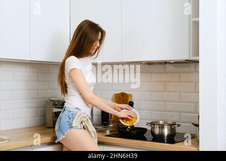 Young woman standing in profile near the electric stove and puts chopped vegetables in a frying pan. Cute woman prepare vegetable gravy for pasta at h Stock Photo