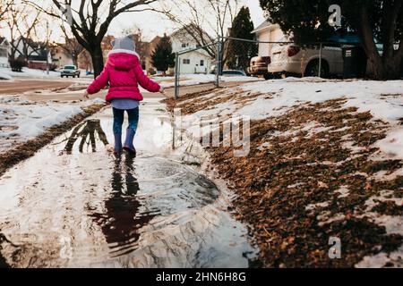 Young girl runs through sidewalk puddle in rubber boots Stock Photo