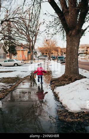 Young girl runs through sidewalk puddle in rubber boots Stock Photo