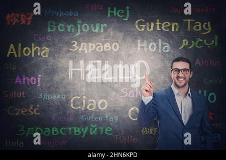 Confident businessman knows different languages. Person shows index finger to the blackboard with word hello translated in multiple foreign languages. Stock Photo