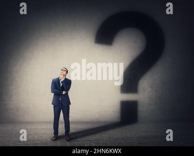 Pensive businessman casting an interrogation mark shadow on the wall. Business questions and decision making concept. Thinking and finding solutions Stock Photo