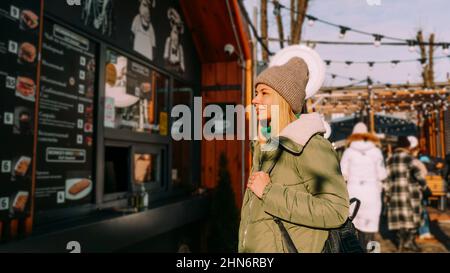 Woman at an urban street food court chooses food from a menu. Street food festival. Sunny day in winter or autumn. The woman is warmly dressed Stock Photo