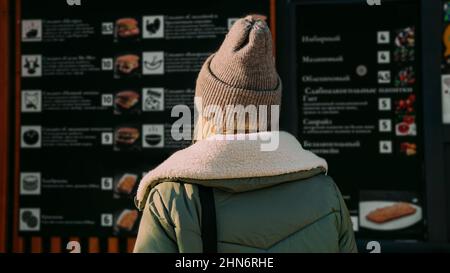 Woman back to the camera at an urban street food court chooses food from a menu. Street food festival. Sunny day in winter or autumn Stock Photo