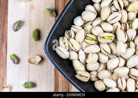 Black square bowl is filled with pistachios.  Bowl is angled on a butcher board background.  Additional pistachios scattered to the left. Stock Photo