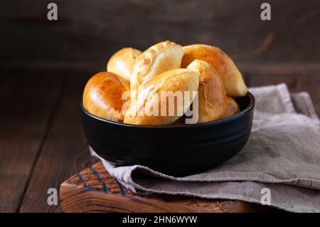 Potato stuffed hand pies - pirozhki in a black bowl on a wooden board with napkin. Stock Photo