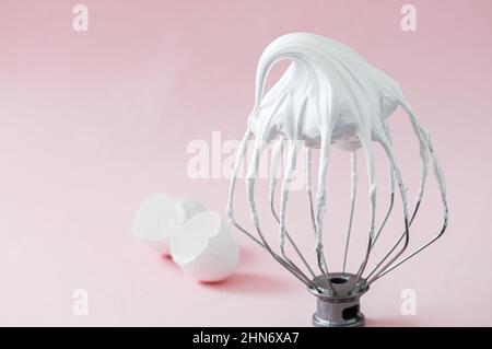Whipped egg whites - beaten italian meringue on a wire whisk and egg shells on pink background. Stock Photo
