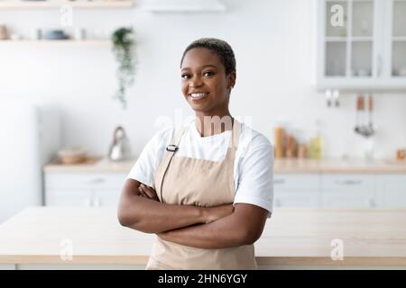 Cheerful millennial african american female in apron with crossed arms looking at camera at modern minimalist kitchen Stock Photo