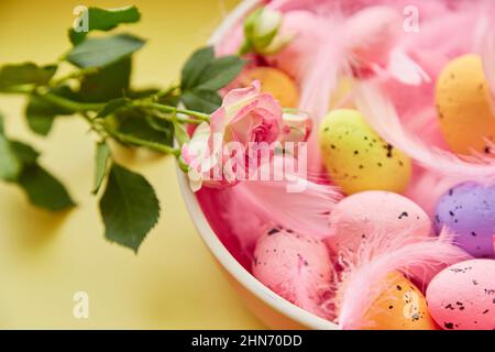Festive Easter background. Pink bowl with colorful eggs, pink feathers and pink rose. Happy Easter concept. Post card on yellow background Stock Photo