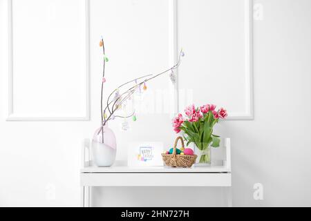 Beautiful Easter composition with basket, painted eggs, flowers and greeting card on table near white wall Stock Photo