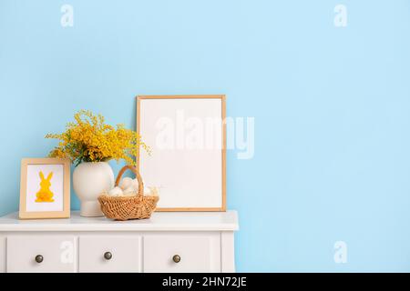 Basket with Easter eggs, mimosa flowers and blank photo frame on table near color wall Stock Photo