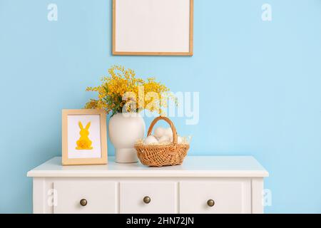 Basket with Easter eggs, mimosa flowers and photo frame on table near color wall Stock Photo