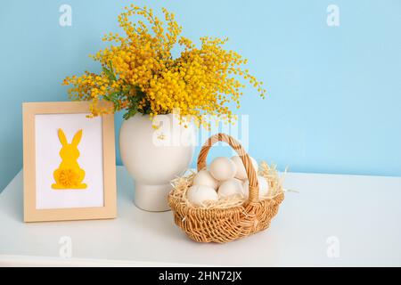 Basket with Easter eggs, photo frame and mimosa flowers on table near color wall Stock Photo