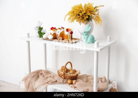 Tray with Easter cakes, eggs and mimosa flowers on shelf unit near white wall Stock Photo
