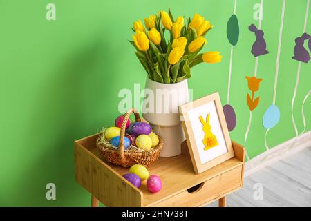 Basket with Easter eggs, photo frame and vase with tulips on table near color wall Stock Photo