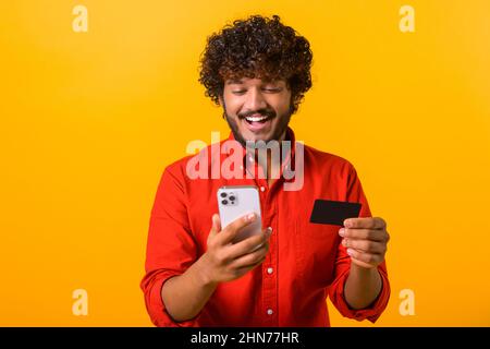 Male paying for long-awaited purchase on the mobile phone while looking at the screen. Middle eastern happy man is ordering food online. Guy is using smartphone and credit card for shopping Stock Photo