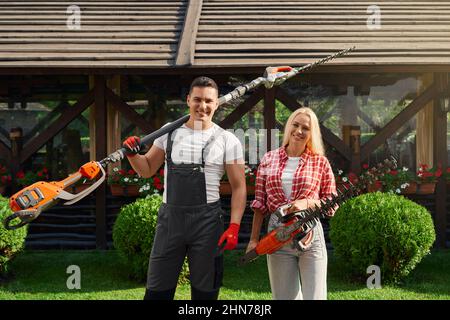 Portrait of happy caucasian man and woman posing at beautiful garden with electric hedge trimmers in hands. Male and female gardeners enjoying seasonal work with plants. Stock Photo