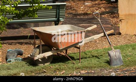 Wheelbarrow filled with cement with shovel leaning on handle and rubber boots near by - work equipment on garden construction site Stock Photo