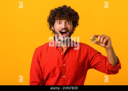 Surprised bearded man holding in his hand golden bitcoin cryptocurrency, looking at camera with astonishment, shocked with price. Indoor studio shot isolated on orange background Stock Photo