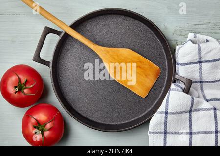 Empty round cast iron pan, wooden spatula, tea towel and tomatoes Stock Photo