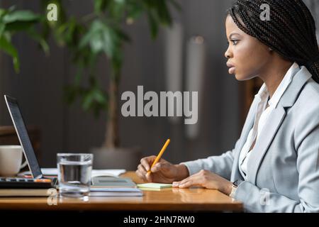 Young black businesswoman concentrated on laptop computer screen writing down notes Stock Photo