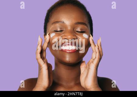 Black african model applying moisturiser face cream to her cheek isolated over purple background. Toothy smile. Facial skincare. Happy face. Stock Photo