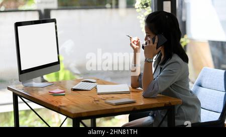 A successful female manager talking on the phone while looking some details on pc desktop computer screen. Stock Photo