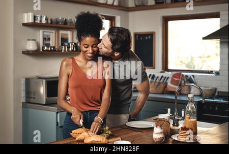 Theres no way Im skipping breakfast with you. Shot of a man kissing his wife while she prepares breakfast in the kitchen at home. Stock Photo