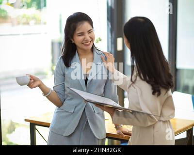 Friendly female officer enjoy talking with her colleague in the office. Happy businesswoman drinking coffee while talking with her coworker. Stock Photo