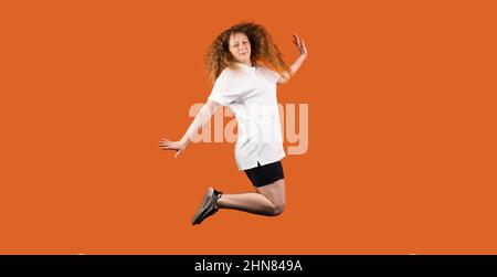 Full-length photo of a fan football redhead woman in white-black uniform jumping with hands by sides on the orange background. Stock Photo