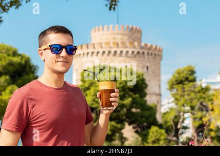 A happy smiling young man drinks traditional Frappe coffee against the background of the famous White Tower in the city of Thessaloniki in Greece. Stock Photo