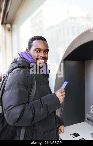 Friendly African American male in outerwear with backpack smiling and looking at camera. while using modern ATM on city street to withdraw money from credit card Stock Photo