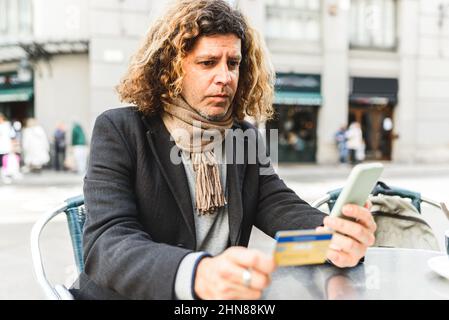 Middle aged man in smart casual outfit with scarf and curly hair. He is using credit card and cellphone to pay for online purchase while sitting at table in street cafeteria Stock Photo
