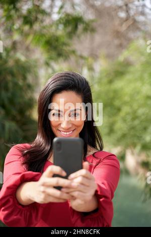 Charming Hispanic female smiling and taking self portrait while standing on street with lush green trees in city on summer day Stock Photo
