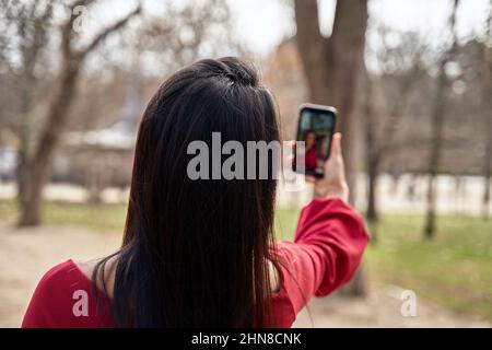 Back view of anonymous female with black hair taking self portrait on smartphone while standing in park with high trees Stock Photo