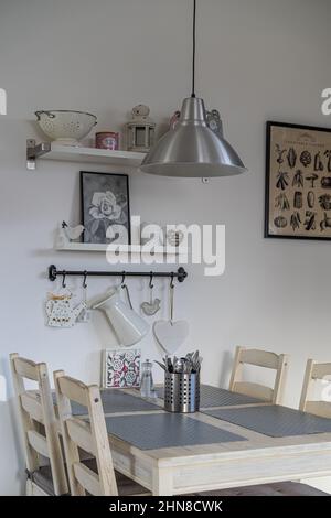 white wall with decorated shelves and wooden table with gray place setting and cutlery, dining area with stainless steel hanging lamp. Stock Photo