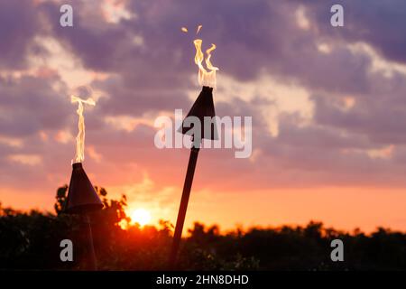 Flames from a pair of tiki torches dance at sunset in front of a colorful Hawaiian sky. Stock Photo