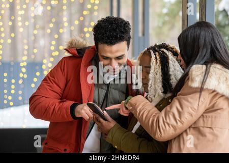 Group of young friends pointing at the screen of mobile phone and smiling while standing on the porch of the house outdoors Stock Photo