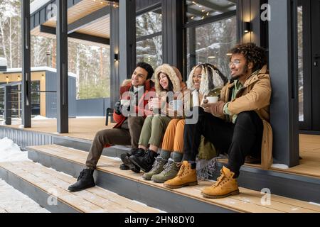 Group of young friends sitting on stairs outside the country house, drinking coffee and enjoying the winter Stock Photo