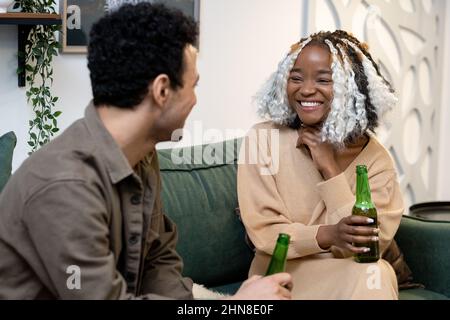 Multiethnic young couple drinking beer and laughing while resting on sofa in the room Stock Photo