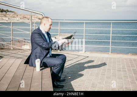 Confident middle age businessman working remotely online, typing on a laptop keyboard while sitting on a beach at sunset. Working remotely on vacation Stock Photo