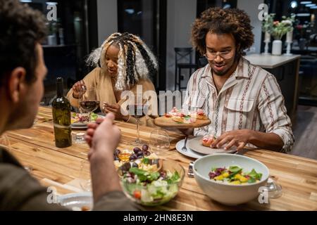 Happy young people sitting at dining table and eating salad and appetizers during dinner party at home Stock Photo