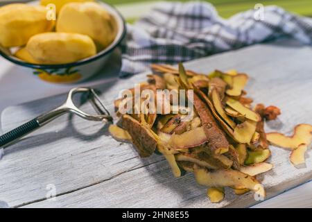 Pile of potato peels on wooden cutting board, food waste for composting. in background, peeled raw potatoes, kitchen towel and  paring knife Stock Photo